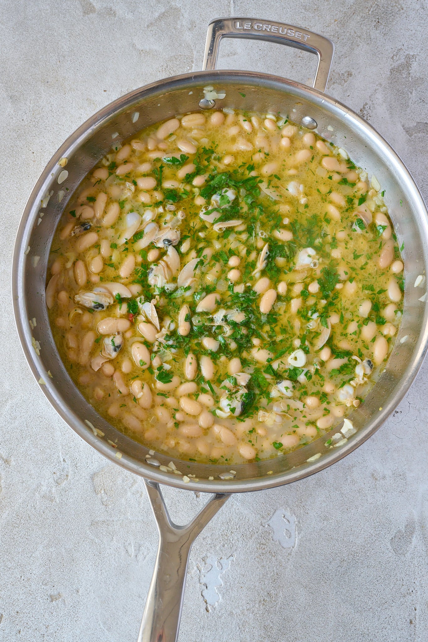 Large stainless steel pan with spanish white bean and clam stew. Fresh parsley is sprinkled all over.