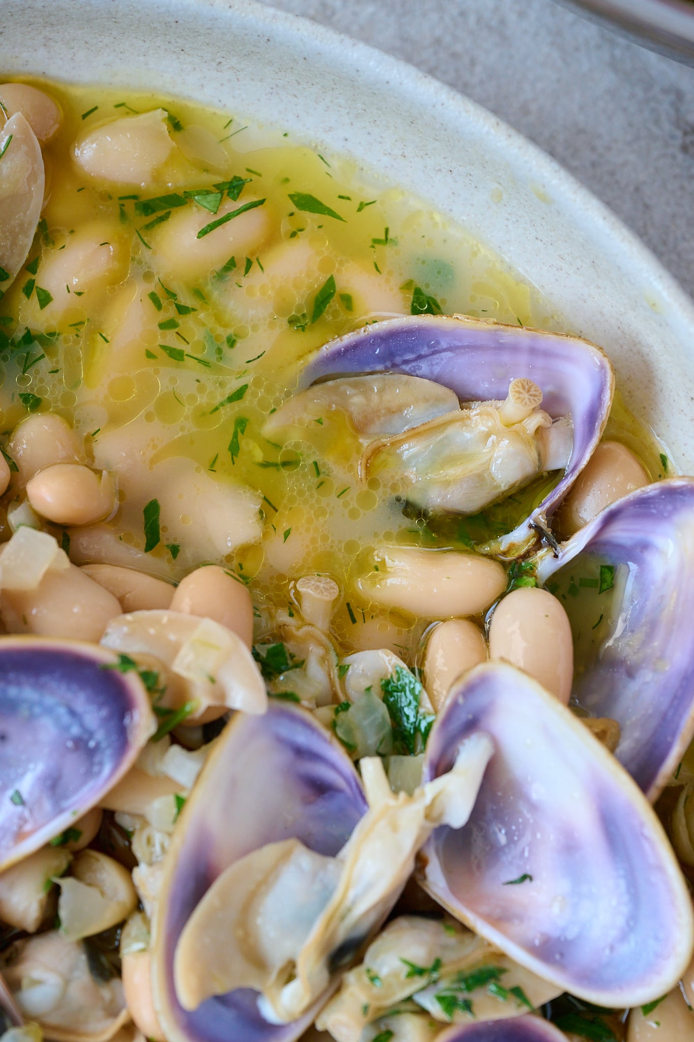 Large white bowl with spanish white bean and clam stew. There are 7 clams that are opened on top with a beautiful purple shell. Fresh parsley is sprinkled all over.