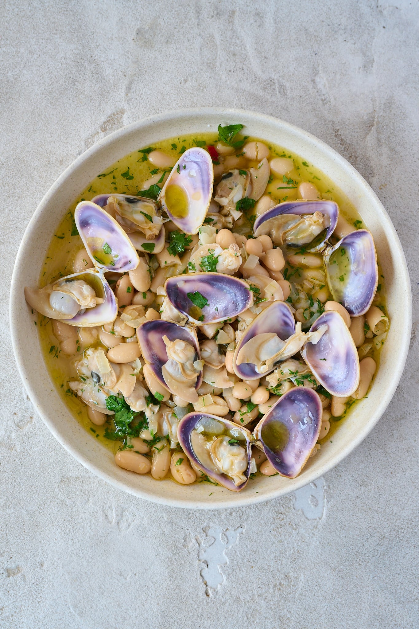 Large white bowl with spanish white bean and clam stew. There are 7 clams that are opened on top with a beautiful purple shell. Fresh parsley is sprinkled all over.