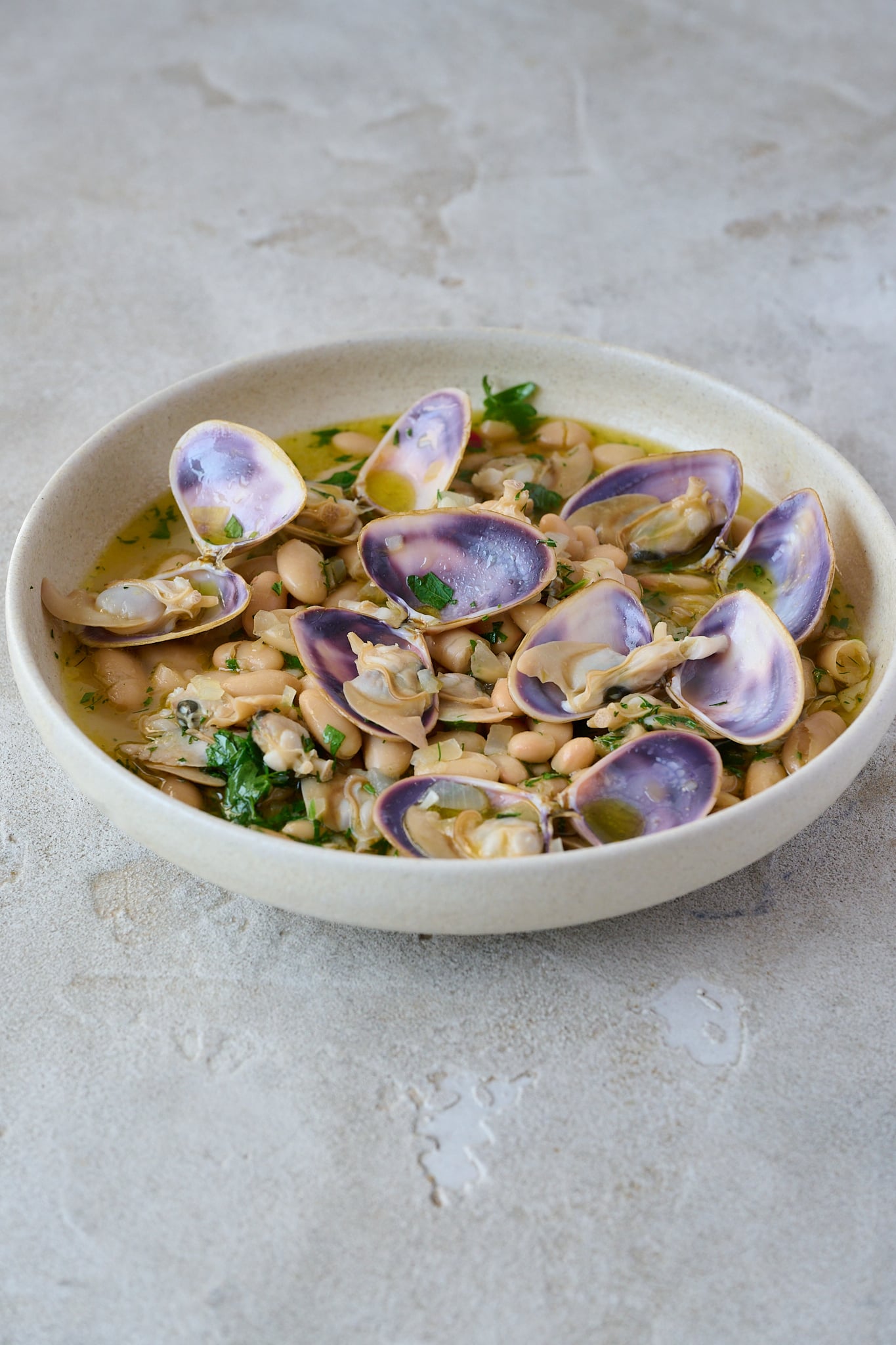 Large white bowl with spanish white bean and clam stew. There are 7 clams that are opened on top with a beautiful purple shell. Fresh parsley is sprinkled all over.