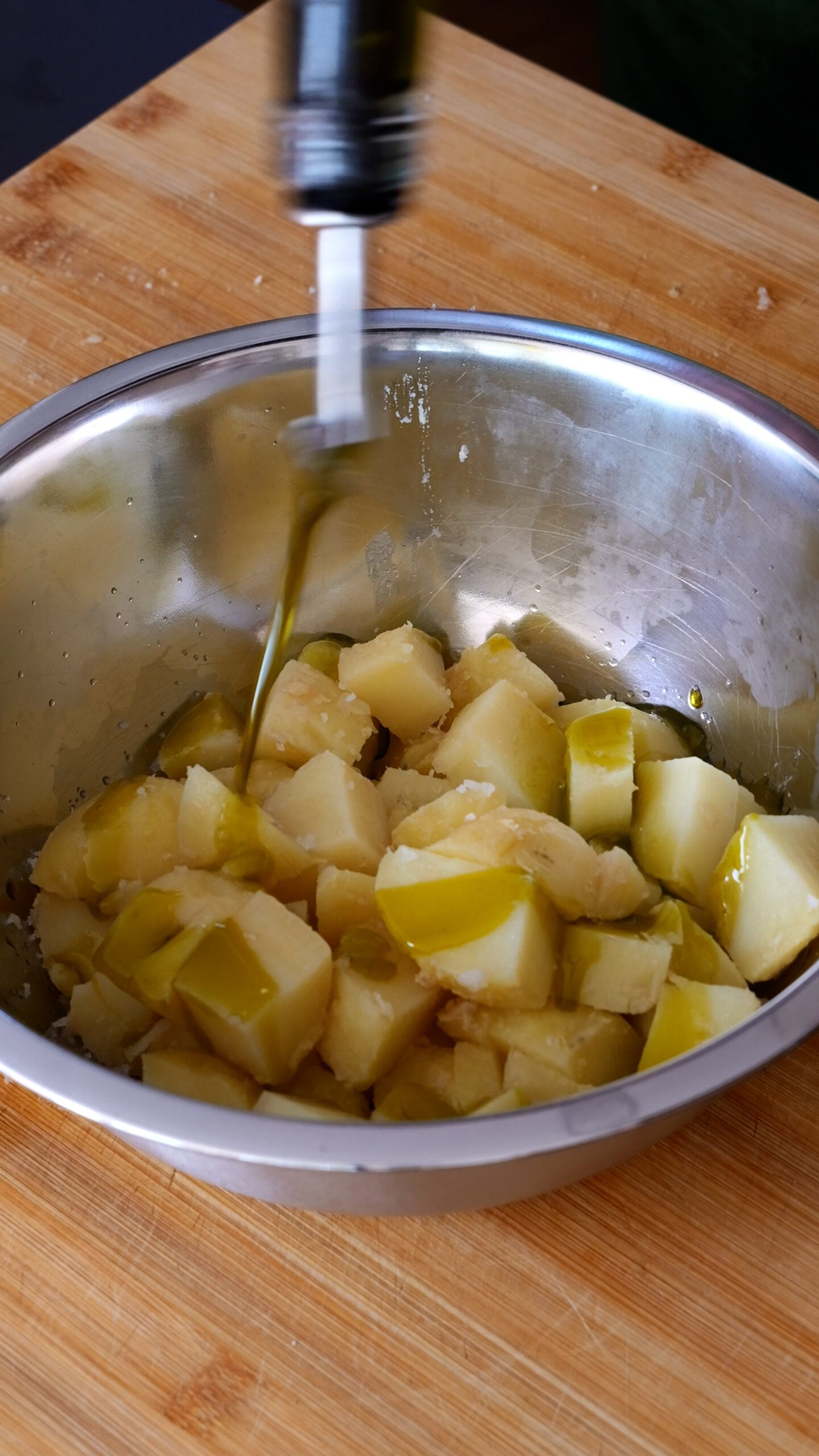 Boiled potatoes are in stainless steel bowl and being dressed with olive oil
