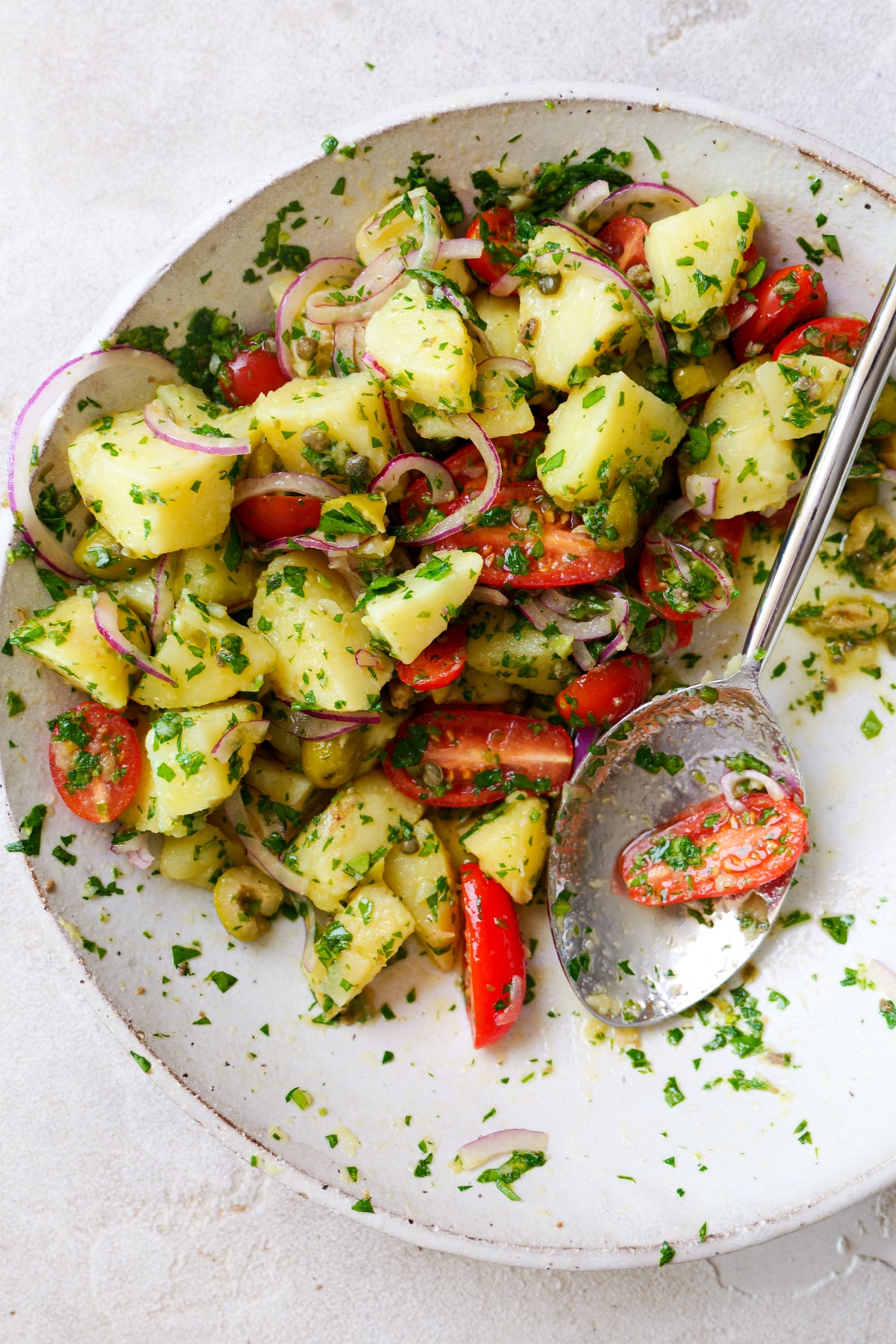 Italian potato salad in large white bowl with lots of fresh parsley sprinkled all over. Large silver spoon is sticking out of right side of bowl with cherry tomato on it.