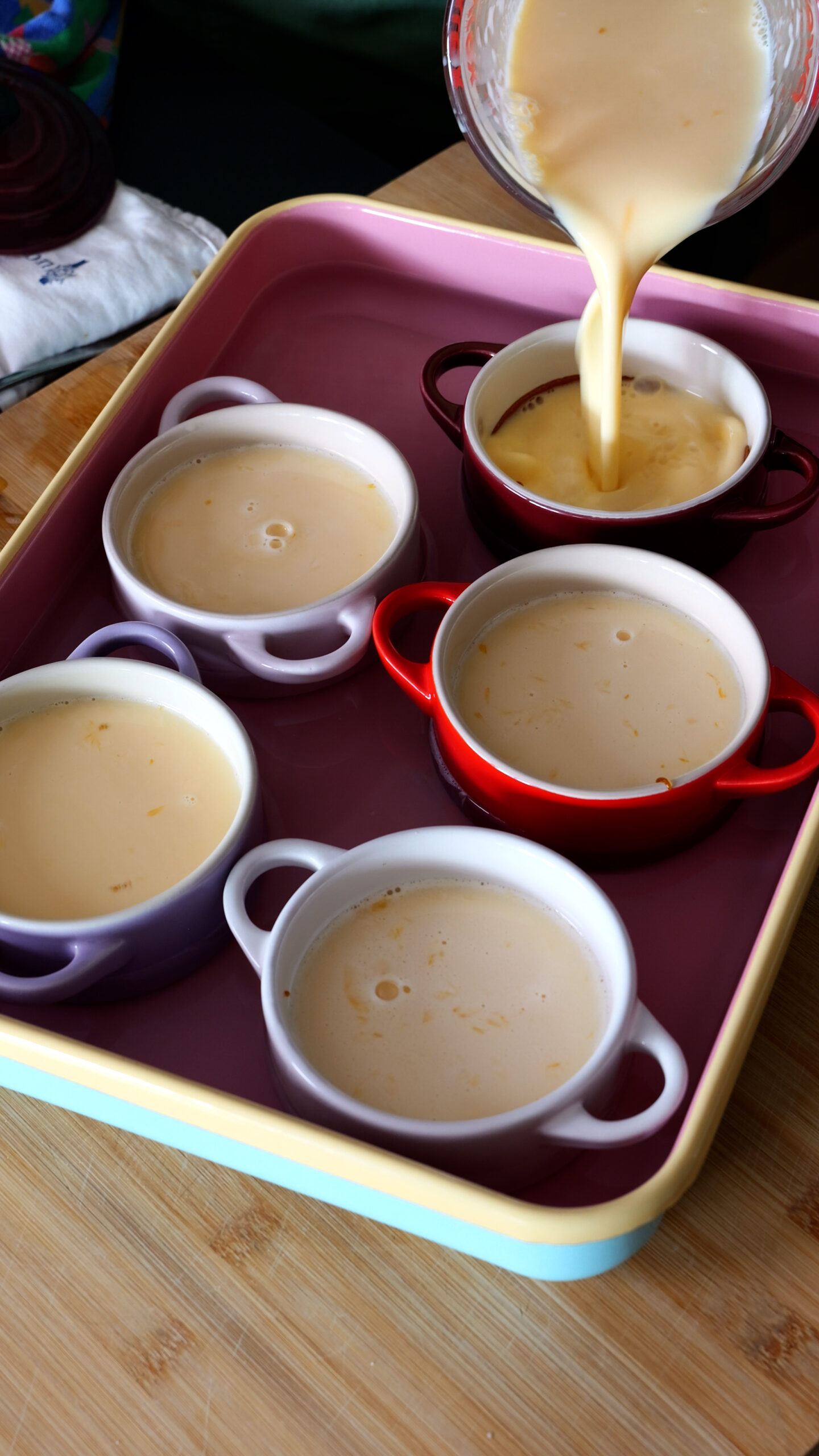 five ramekins are in large pink baking tray and being filled with custard.