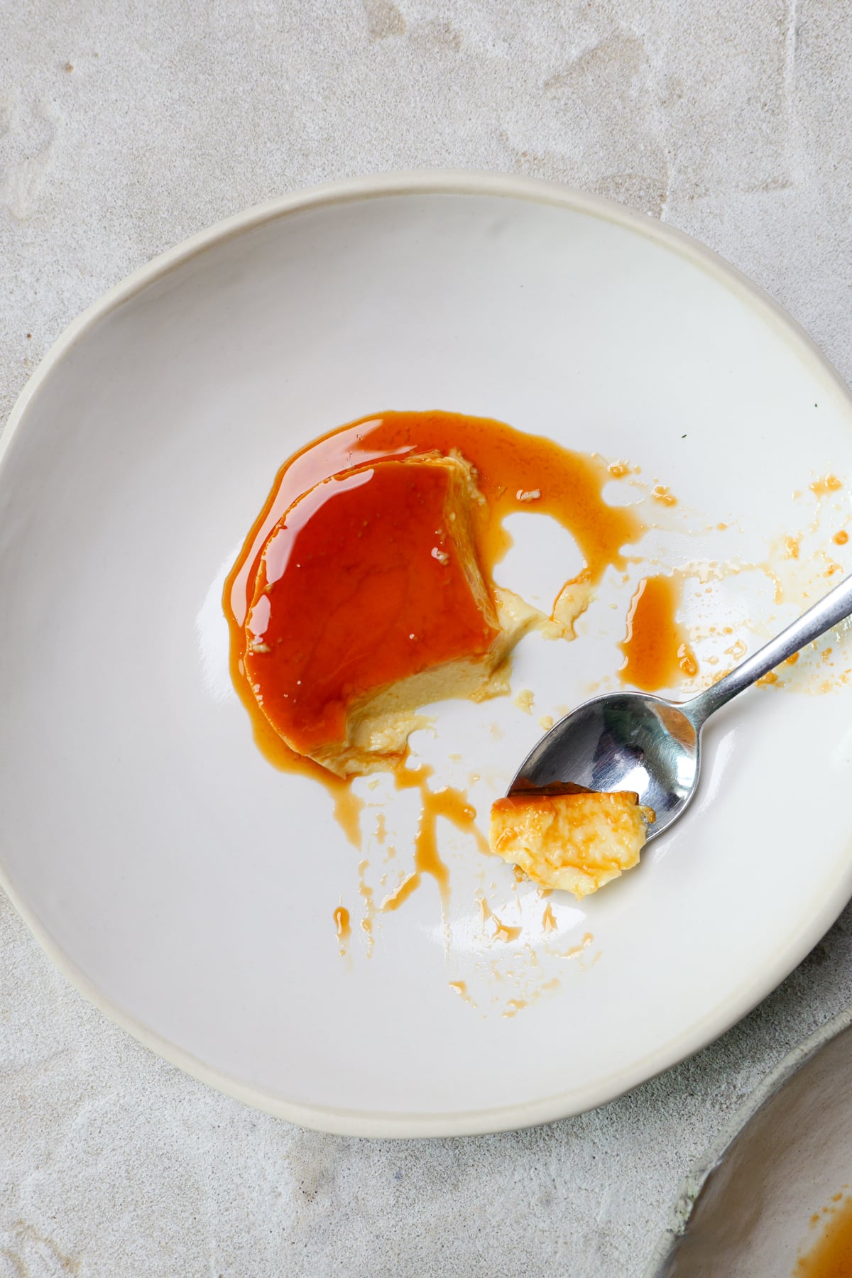 Spanish flan is in large white bowl with golden custard drizzled all over. A silver spoon has broken into the flan.